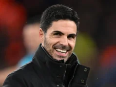 Julian Alvarez entra na foto de transferência do Arsenal Portrait of Mikel Arteta smiling, wearing a black jacket against a blurred stadium background.