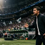 Treinador de futebol na área técnica, à beira do campo, durante partida noturna com estádio lotado ao fundo.